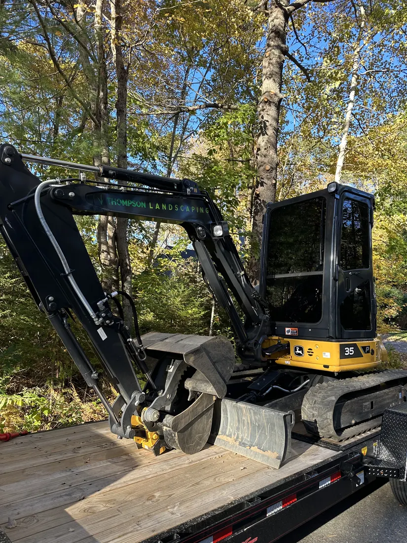 Compact excavator on a trailer beside a wooded area